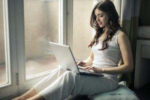 woman wearing tank top sitting by the window looking at laptop screen scaled